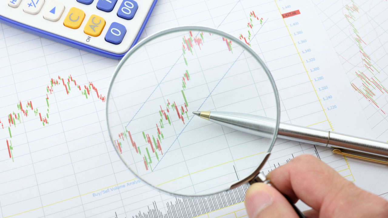 Hand holding a magnifying glass over a financial chart with candlestick patterns, analyzing trading signals for market trends. What Are Trading Signals & How Do They Work?