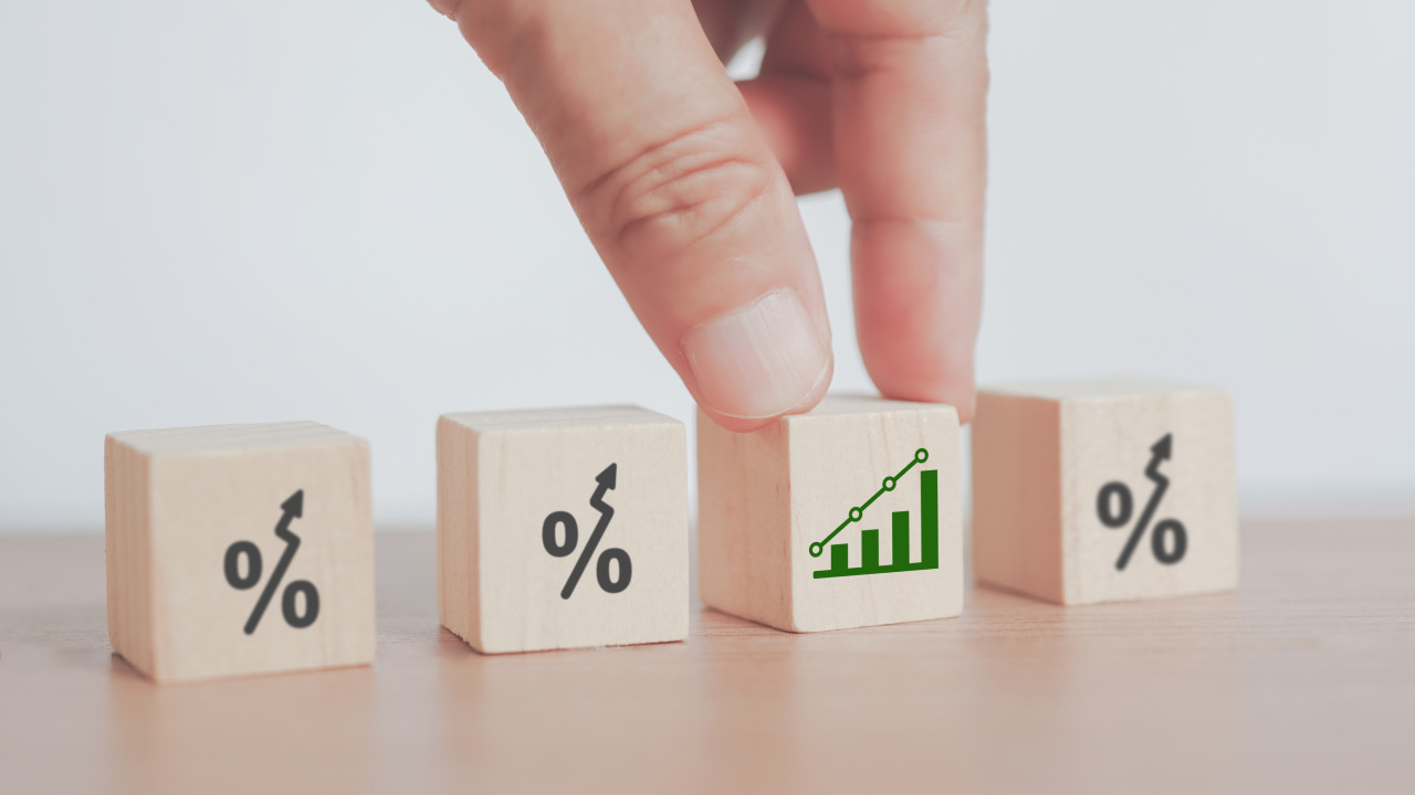 A hand picks up a wooden block displaying a green upward-trending bar chart, positioned between three other blocks that show percentage signs with upward arrows.