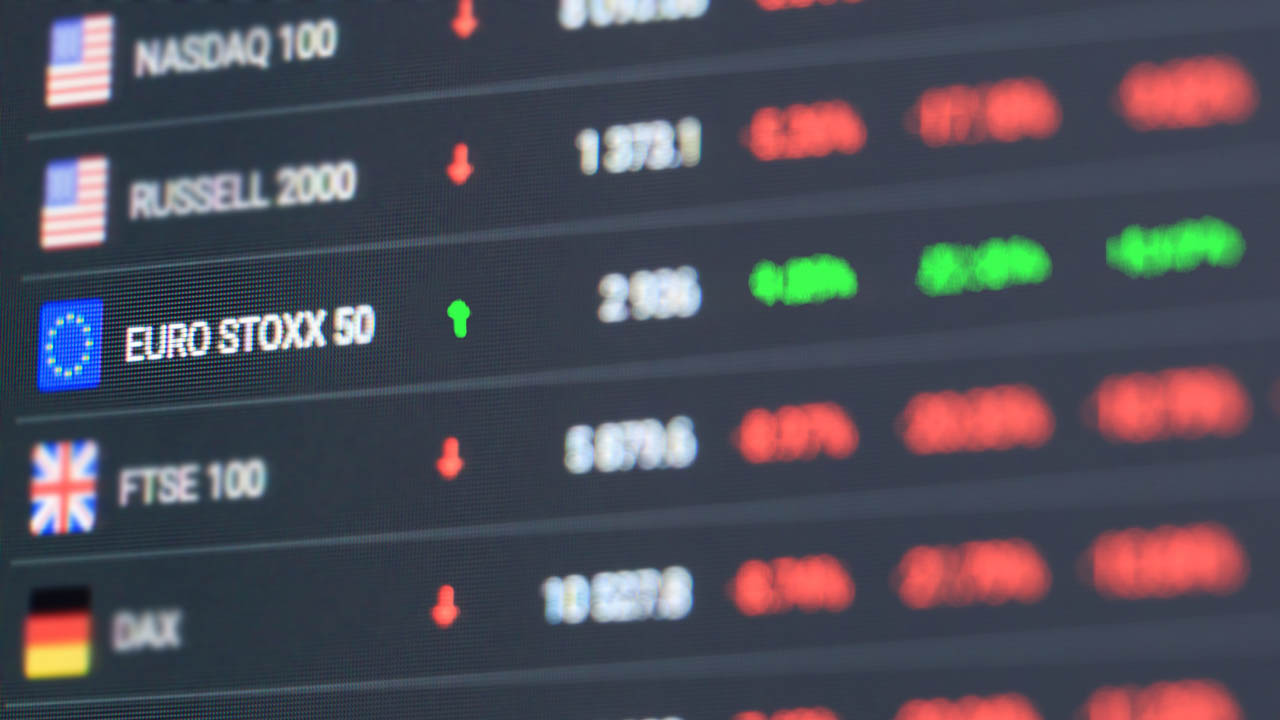 A digital trading screen displaying stock indices, including the Euro Stoxx 50, Nasdaq 100, FTSE 100, and DAX, with green and red indicators showing market movements.