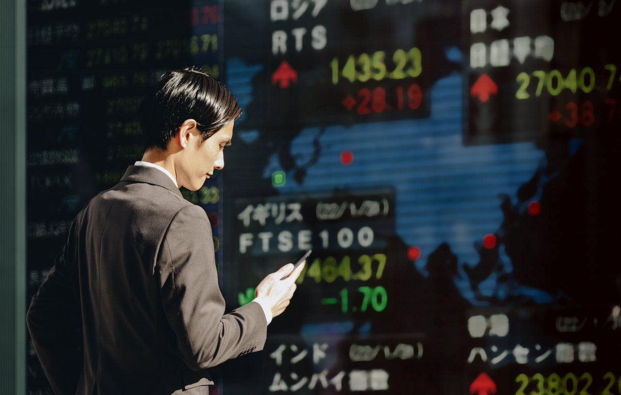 Japanese Markets to Rebound After its Biggest Drop in August's First Week — Businessman in a suit checking his phone in front of an electronic stock market board displaying Japanese and international indices, highlighting the rebound of Japan's equities.