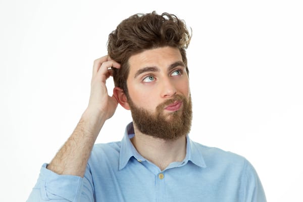 Man with a beard scratching his head and looking confused or thoughtful, wearing a light blue shirt against a white background.