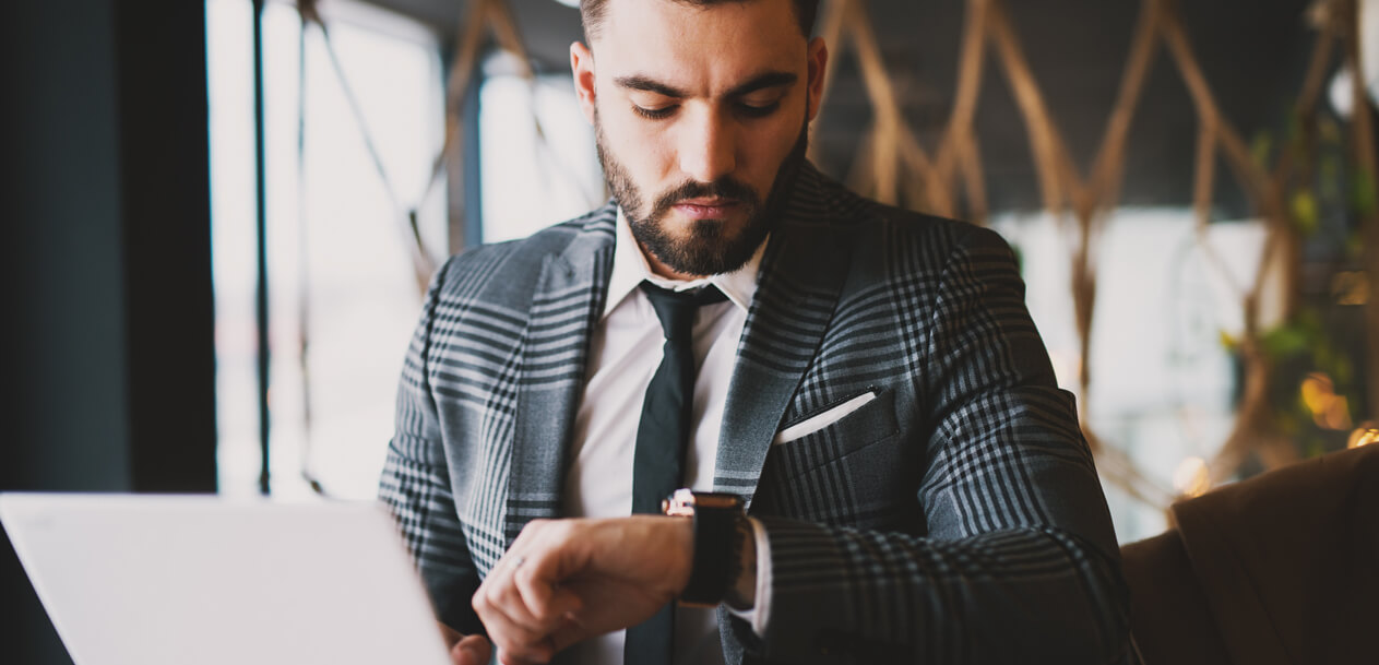 The Best Time To Trade Forex, illustrated by a man in a checkered suit looking at his watch while working on a laptop in a stylish indoor setting.