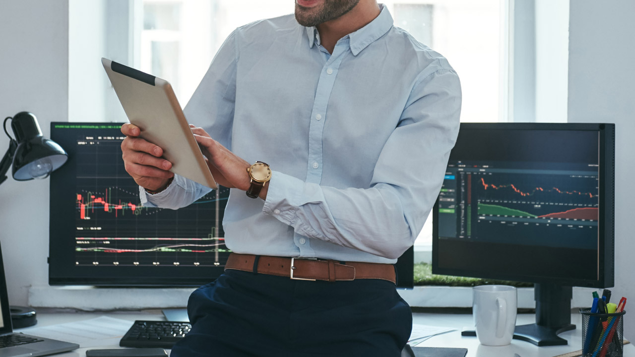 Effective Risk Management in Crypto Trading – A trader in a professional setting analyzing market data on a tablet, with multiple monitors displaying financial charts and trading indicators in the background.