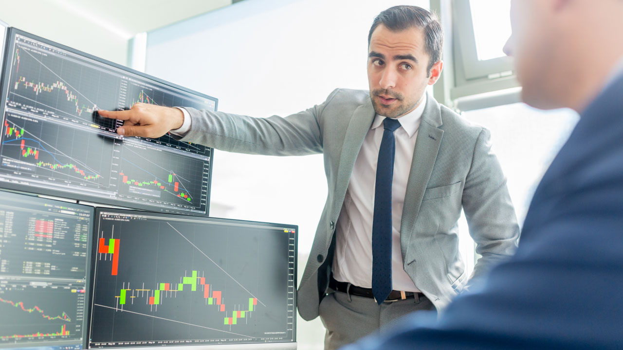 Why do forex brokers charge commission fees? A financial analyst in a suit explains forex charts on multiple monitors, highlighting trading costs and broker fees in a professional office setting.