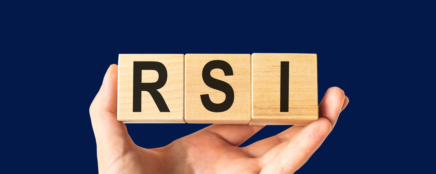 Trading With The RSI Indicator, illustrated by a hand holding three wooden blocks spelling out 'RSI' against a dark blue background, representing the RSI trading indicator.