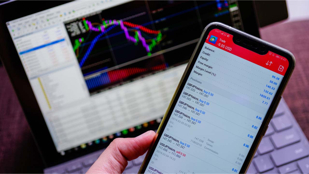 How Does MetaTrader 4 Work? A trader using the MetaTrader 4 platform on a smartphone, displaying open forex positions, with a trading chart on a laptop in the background.