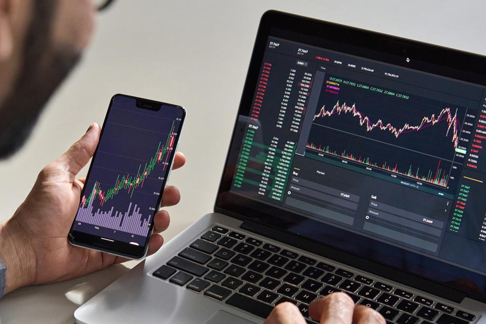Trader analyzing financial charts on a smartphone and a laptop simultaneously, illustrating the difference between scalping and swing trading strategies.