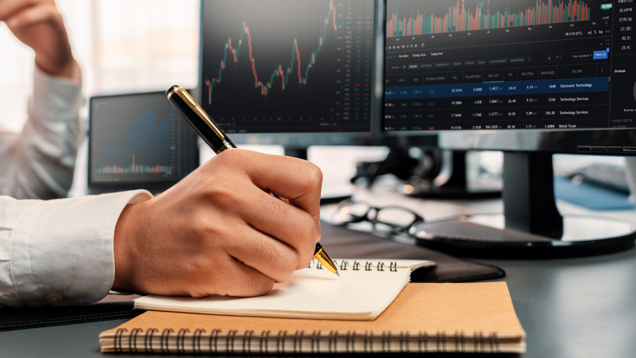 a person’s hand writing in a spiral notebook with a gold pen, seated at a desk with multiple monitors displaying financial trading charts and stock market data