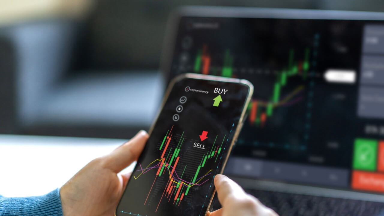 Trader holding a smartphone displaying a forex trading app with buy and sell indicators, analyzing market charts with a laptop in the background. Top 10 Essential MT4 Indicators for Technical Analysis.