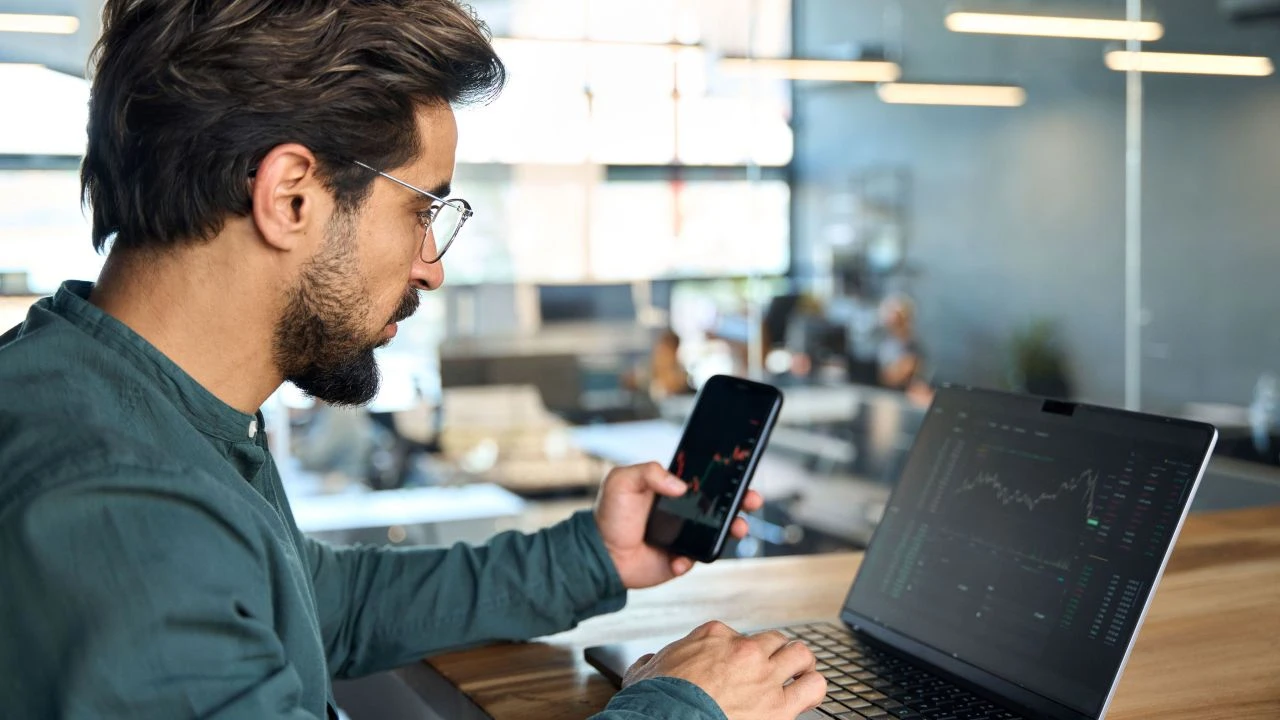 Trader holding a phone in front of a laptop, analyzing forex charts to understand the ABCD pattern in forex trading.