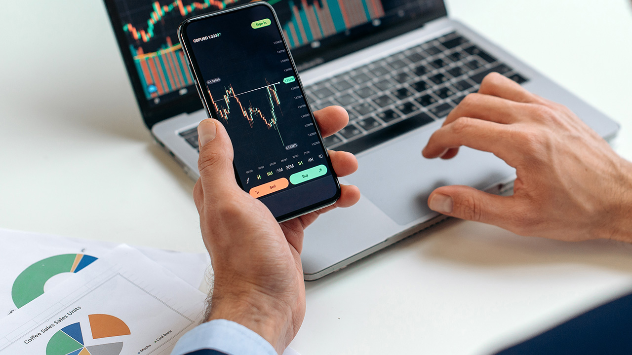 Trader using a smartphone and laptop to analyze financial charts, illustrating why use custom moving averages to tailor your strategy.