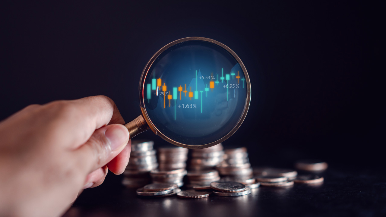 Hand holding a magnifying glass focused on a candlestick chart with percentage gains, placed over stacked coins, illustrating why forex traders should use a risk calculator.