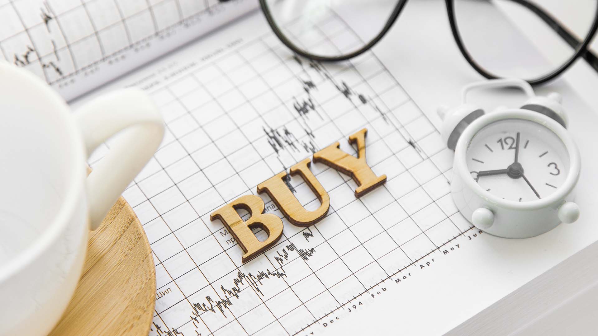 Buy and Hold: The Long-Term Trading Strategy – A wooden 'BUY' sign placed on a financial chart with a small clock, glasses, and a coffee cup, symbolizing long-term investment and market analysis.