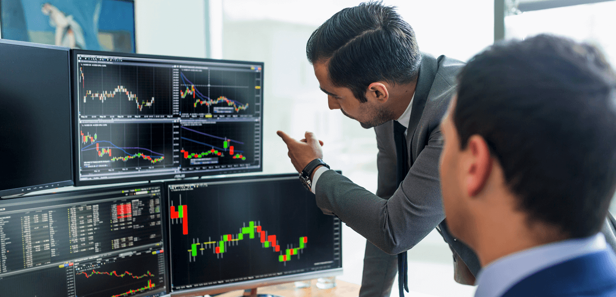 Two traders in formal business attire analyzing multiple forex trading charts on computer monitors, discussing market trends and potential strategies.