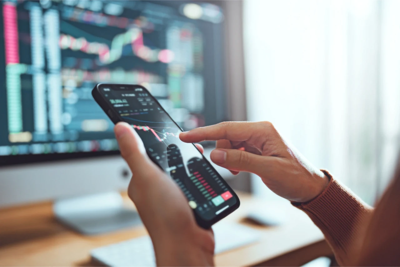 A person using a smartphone to analyze stock market charts, with a trading dashboard visible on a computer monitor in the background, illustrating why retail traders are often wrong.