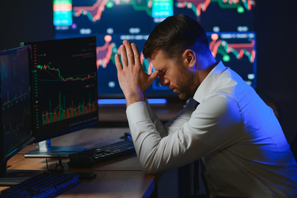 astressed trader sits at his desk with his hands on his forehead, surrounded by multiple screens displaying market charts, representing the challenges of handling trading slumps.