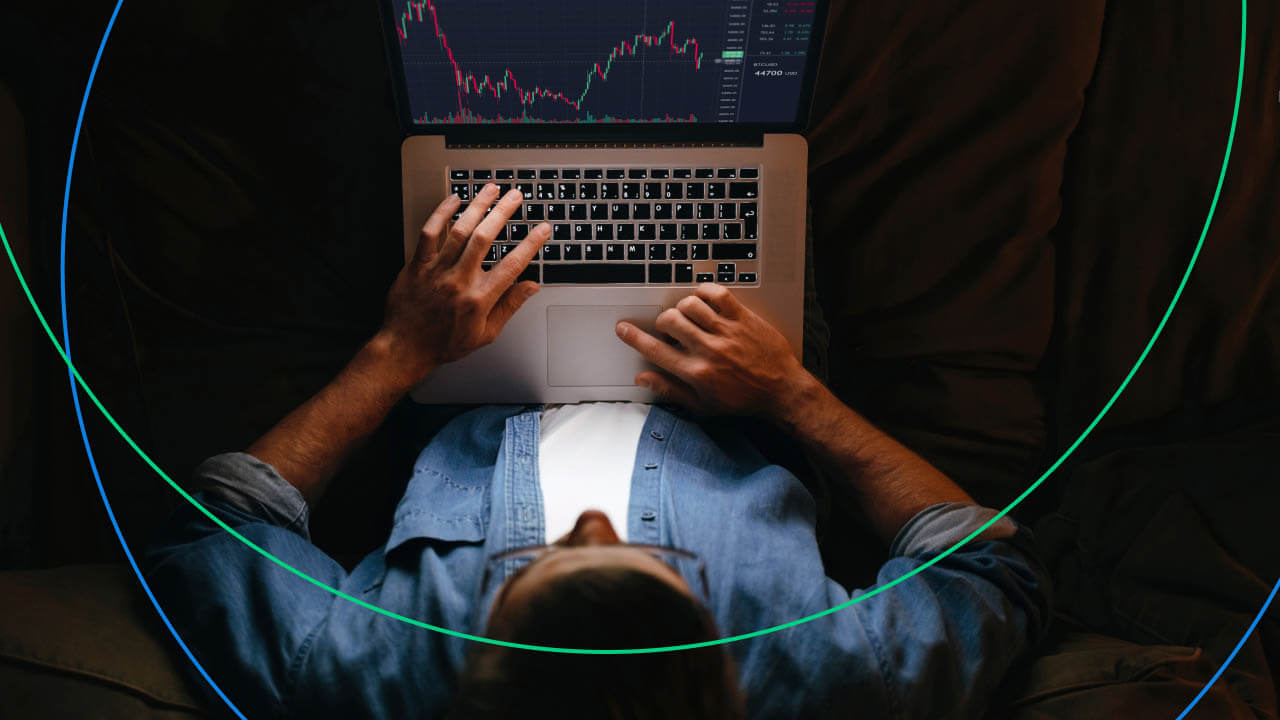 a person wearing a denim shirt is seen from above, sitting comfortably and using a laptop to monitor financial charts