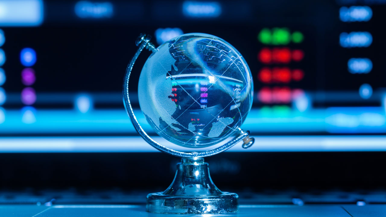 A crystal globe on a stand placed in front of a blurred digital screen displaying market data