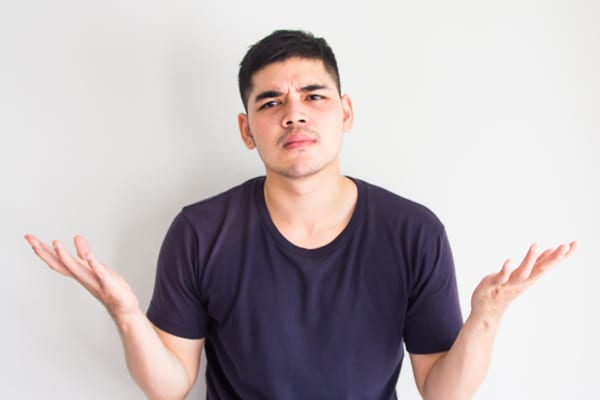 Confused man shrugging with both hands raised, wearing a dark t-shirt and standing against a plain light-colored background.