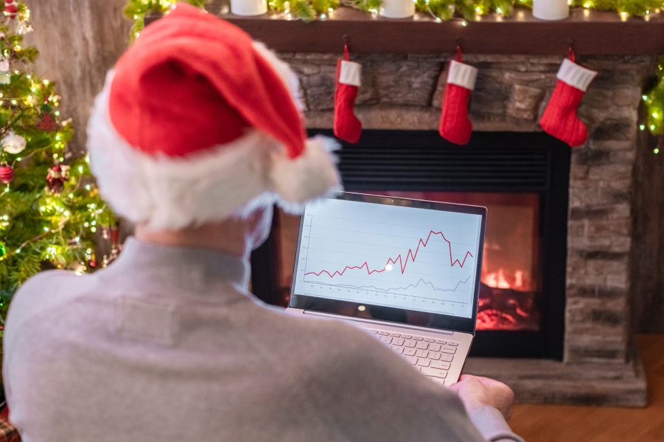 A trader wearing a Santa hat analyzing a forex chart on a laptop by a cozy fireplace with Christmas decorations. Assessing forex market activity during the holiday season.