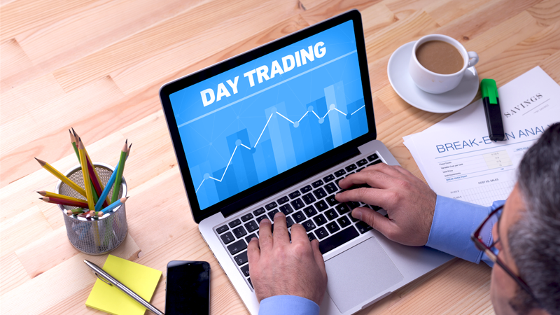 Person typing on a laptop displaying "DAY TRADING" on the screen with a blue background and a rising chart line.