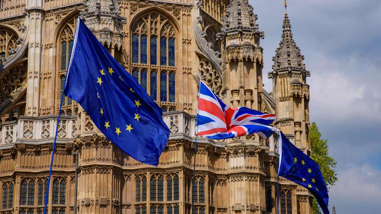 European Union flags and a Union Jack (United Kingdom flag) fly in front of a historic Gothic-style building, likely part of the UK Parliament in London.