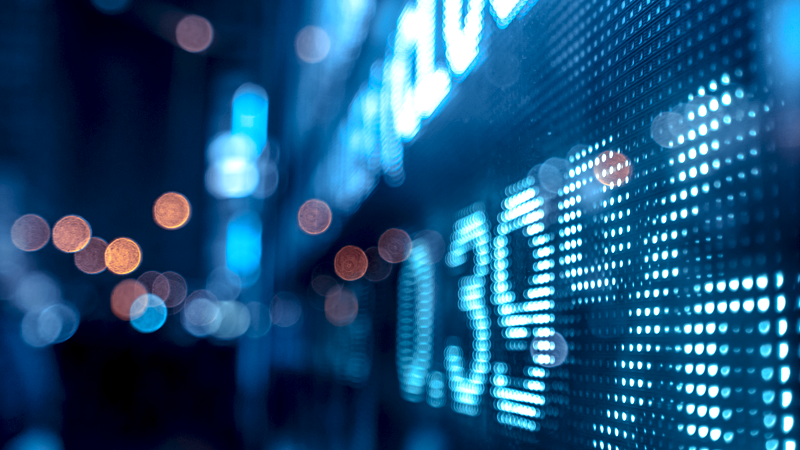 Close-up of a digital stock market display showing numbers and financial data in blue tones, with blurred city lights in the background.