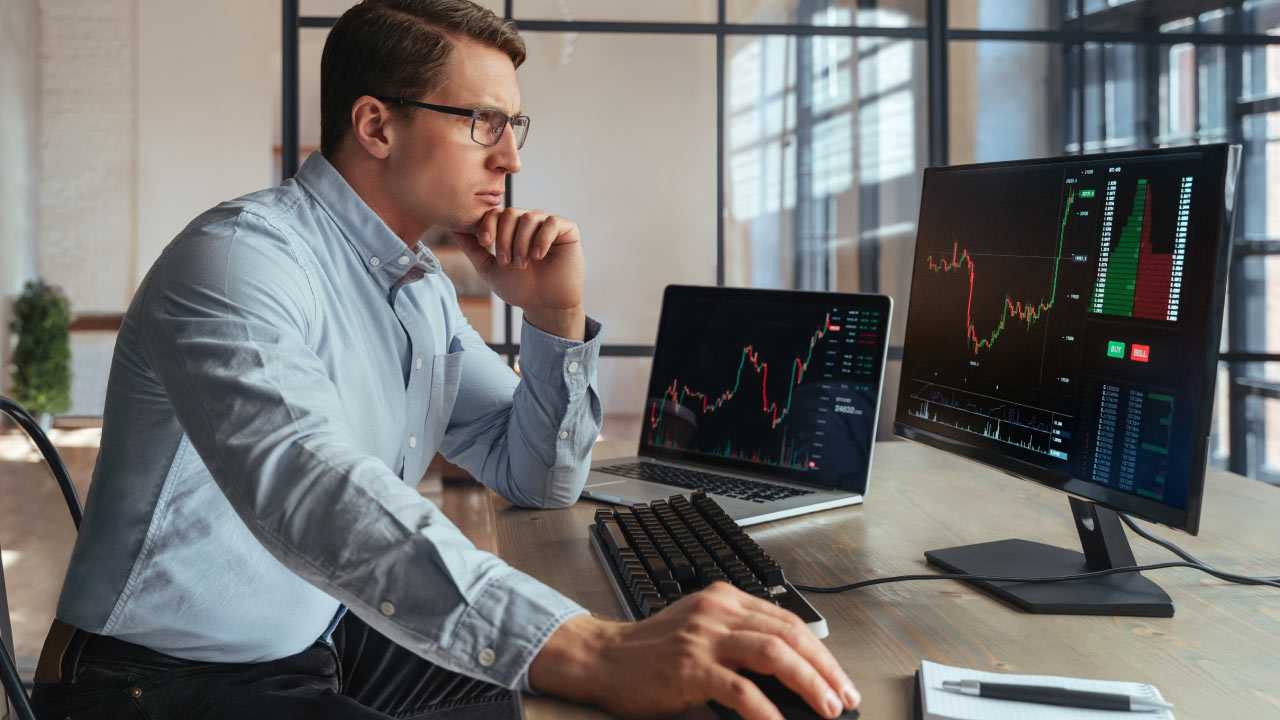 A focused man wearing glasses and a light blue shirt is analyzing financial charts on two monitors at a modern office desk