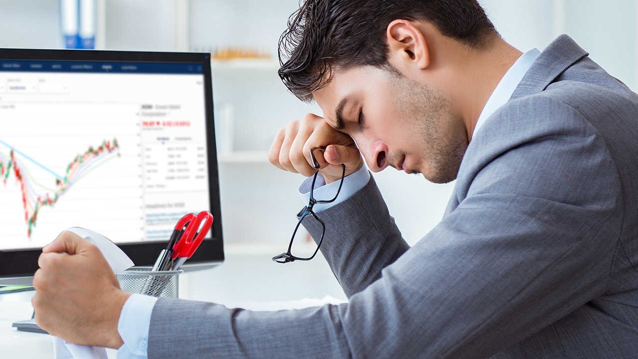 Managing Emotions in High-Stakes Trading — illustrated by a stressed businessman in a suit sitting at his desk, holding his glasses and resting his head on his hand, with a trading chart showing losses displayed on his computer screen in the background.
