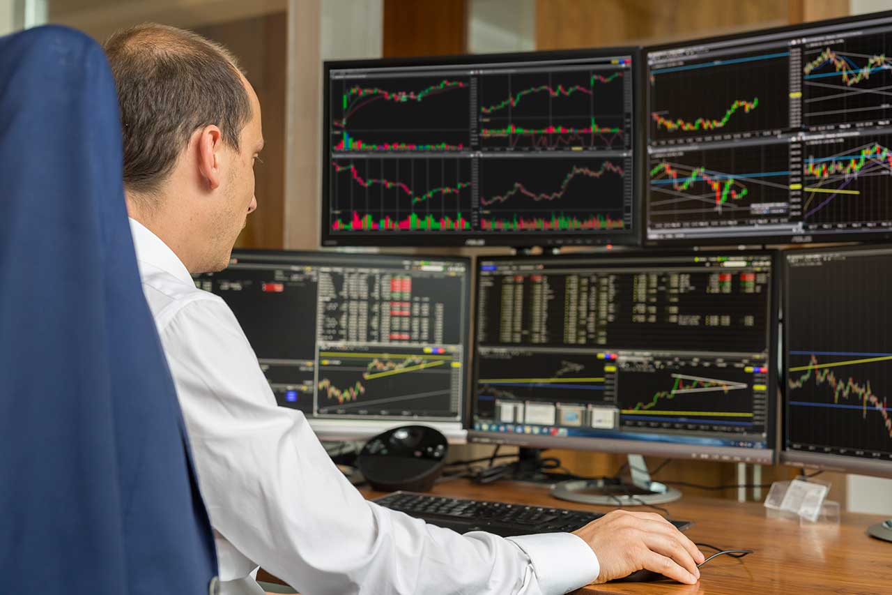 A retail trader in a white shirt analyzing multiple financial charts on six monitors, tracking market trends and price movements in a trading setup.