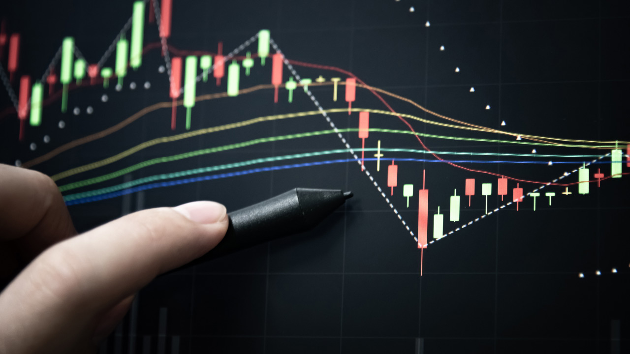 close-up image of a hand holding a stylus pointing at a candlestick chart on a digital trading screen