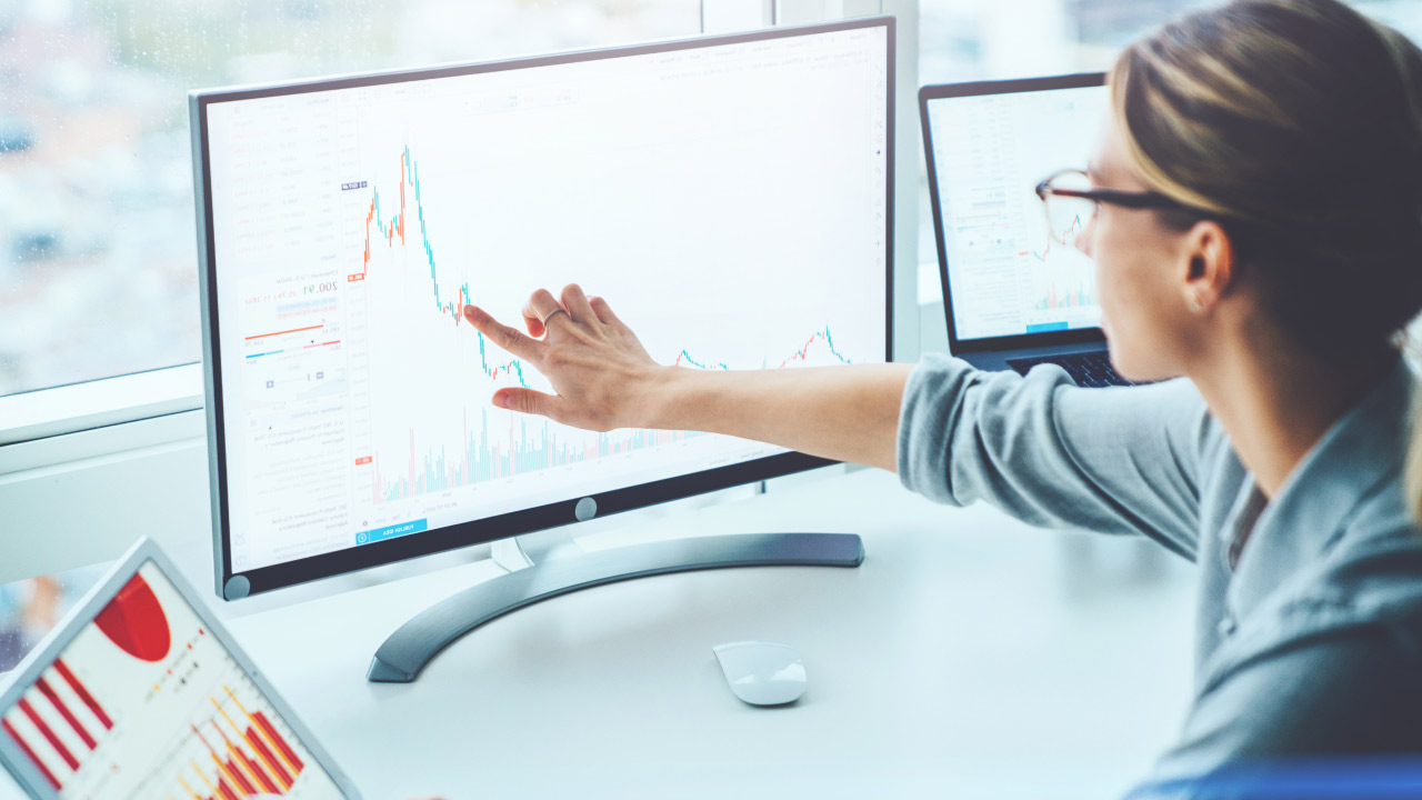 Businesswoman analyzing a stock chart on a large computer monitor using linear regression tools, with additional data displayed on a tablet and a second screen, illustrating the concept of the Linear Regression Slope.