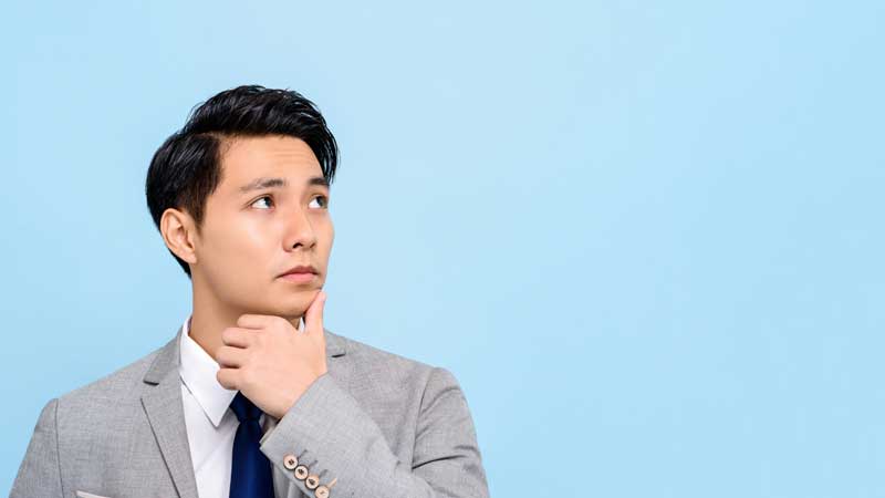 Young man in a gray suit thinking while looking up against a light blue background.