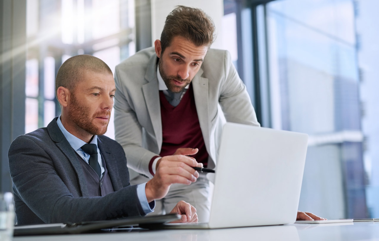 Questions to Ask When Trading Trends, illustrated by two businessmen in suits discussing in an office, with one pointing at a laptop screen with a pen.