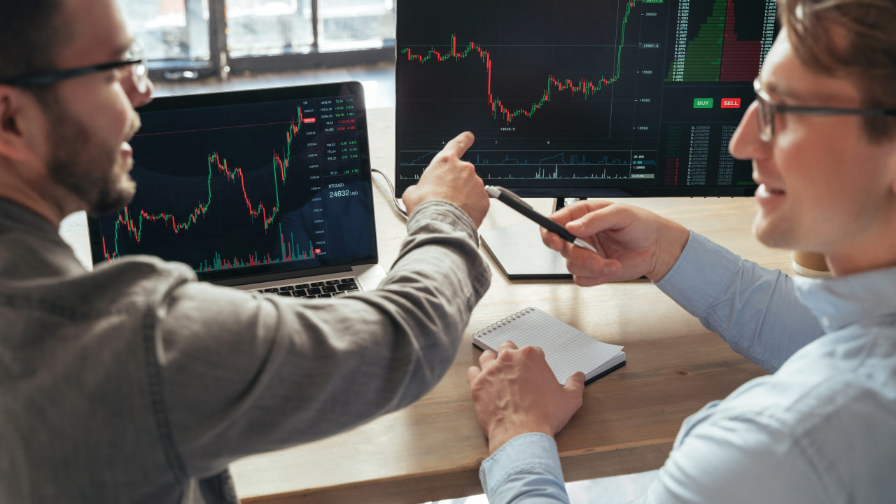Passive vs Active Trading: What's the Difference? – Two traders discussing financial charts displayed on multiple screens, analyzing market trends and strategies.