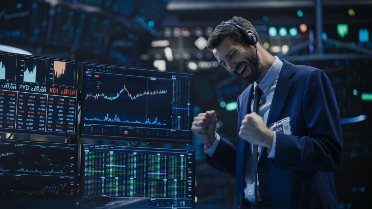A happy trader in a suit celebrates a big win in front of multiple monitors displaying financial charts and data.