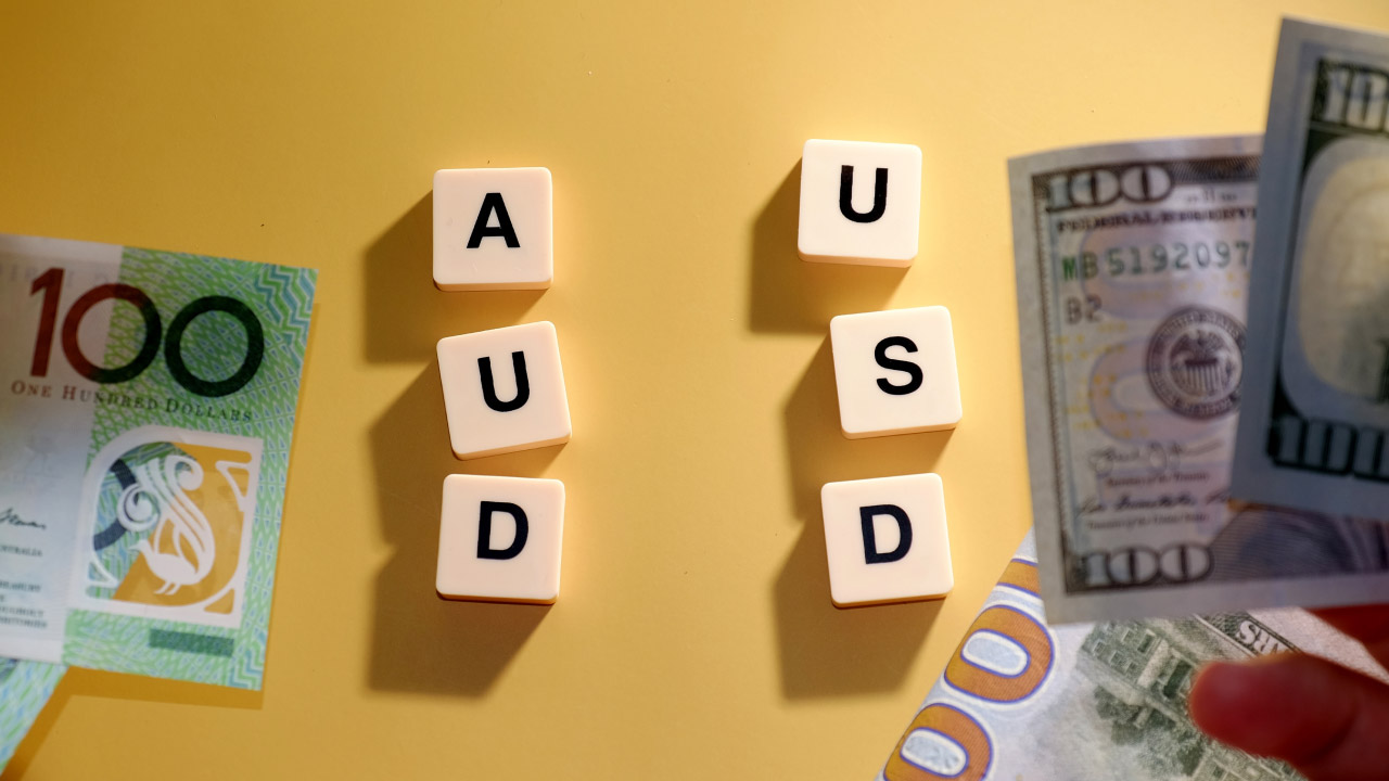 A visual representation of the AUD/USD currency pair. On a yellow background, the letters "AUD" and "USD" are arranged vertically using white letter tiles.