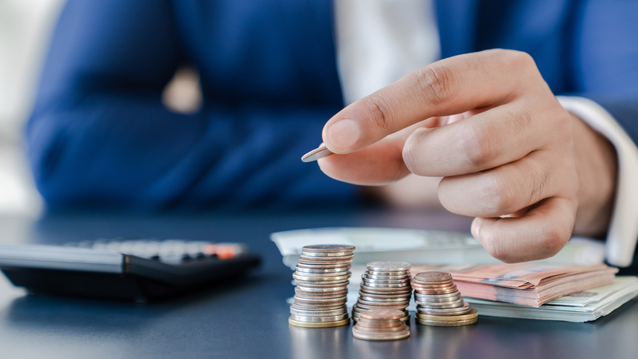 Businessperson stacking coins with cash and a calculator in the background, symbolizing financial planning and setting realistic trading goals.