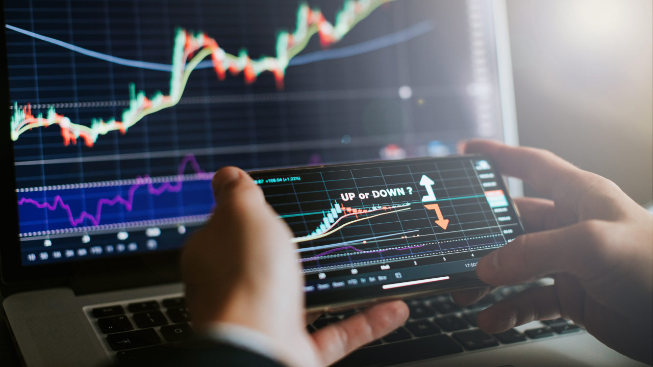 A trader analyzing forex charts on a smartphone and laptop, with 'UP or DOWN?' displayed on the screen, symbolizing the challenge of predicting market movements.