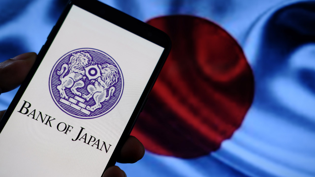 A smartphone screen displaying the Bank of Japan logo held in front of a blurred Japanese flag, illustrating why the BoJ intervenes in the forex markets.