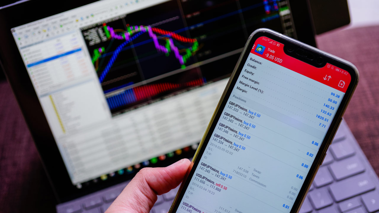 A trader holding a smartphone displaying an MT5 trading account with active positions, while a laptop screen in the background shows the MetaTrader 5 platform with trading charts and indicators. Learn How to Place a Trade on MT5 efficiently.