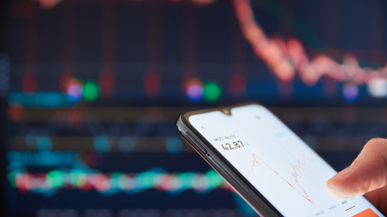 A trader monitors price movements on a smartphone with a blurred trading chart in the background, illustrating how to set up the perfect trading routine.